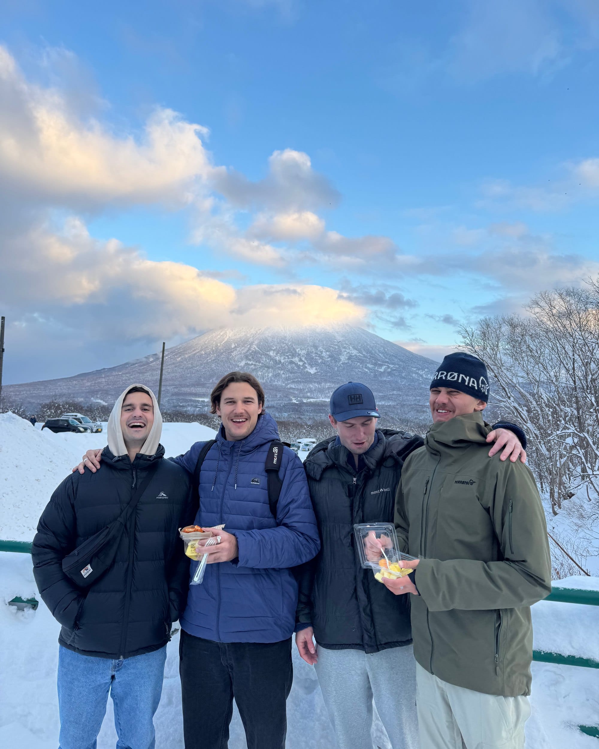 Four friends in winter jackets laughing together in the snow, a snow-capped mountain behind them