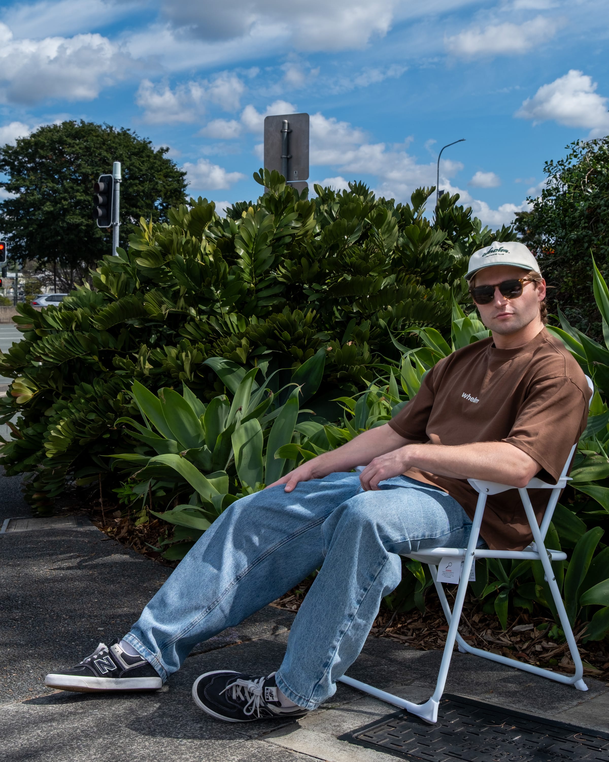 Model in the whelm Deep Mocha Tee and Two Tone Cap, sitting in a white folding chair on a footpath